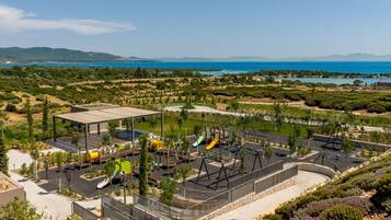 A playground with various play equipment, a covered seating area, and a view of the sea and hills in the background.