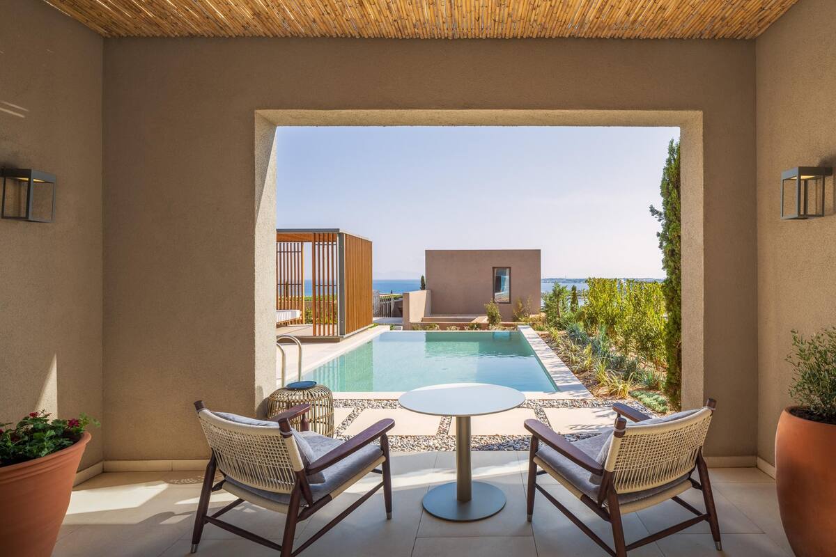 Area piscina con tavolo e sedie, vista sul mare e un edificio sullo sfondo.