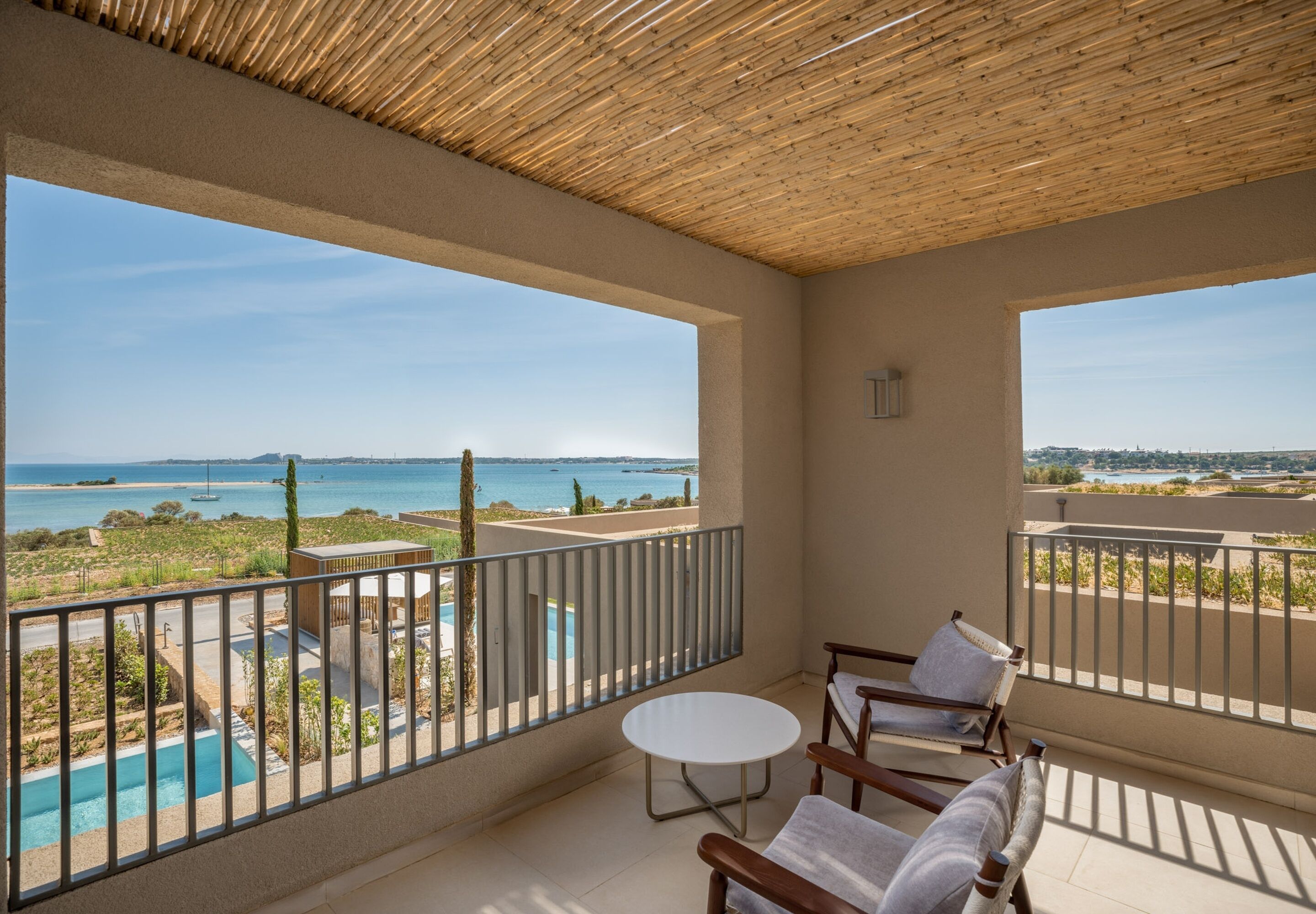 A balcony with a view of a pool and the sea, wooden furniture, and a thatched ceiling.
