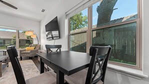 A dining area with a table and chairs, a flat-screen TV mounted on the wall, and large windows with a view of the outdoors.