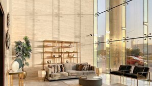 A spacious living room with a large sectional sofa, a round coffee table, and a glass wall offering a city view. - Modern JVC Studio with Pool Gym and Easy Access to Attractions (Dubai)