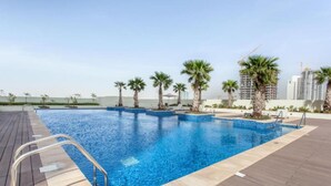 A swimming pool with a wooden deck, surrounded by palm trees and modern buildings in the background. - Modern JVC Studio with Pool Gym and Easy Access to Attractions (Dubai)