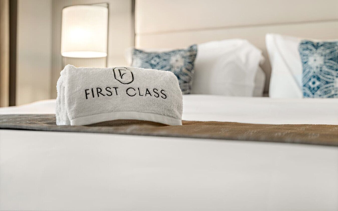 A neatly made bed with a white towel on it that reads "FIRST CLASS".