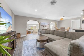 A spacious living room with a large sectional sofa, a coffee table, and a TV on a wooden console. There are two archways leading to another room, and a view of the ocean through a large window. - 3BR home w/ pool & BBQ grill (Satellite Beach)