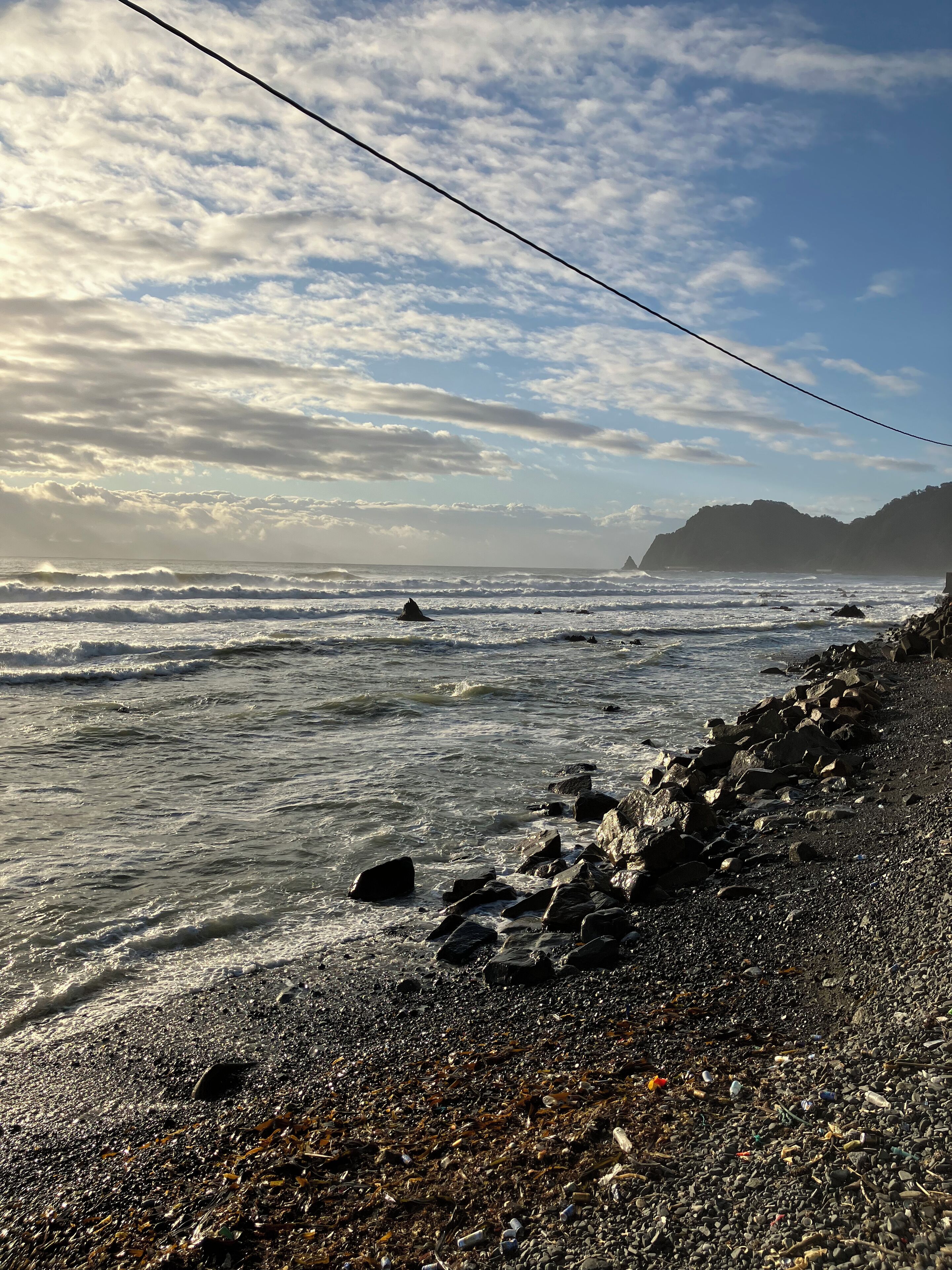 A rocky beach with waves, a power line, and distant hills.