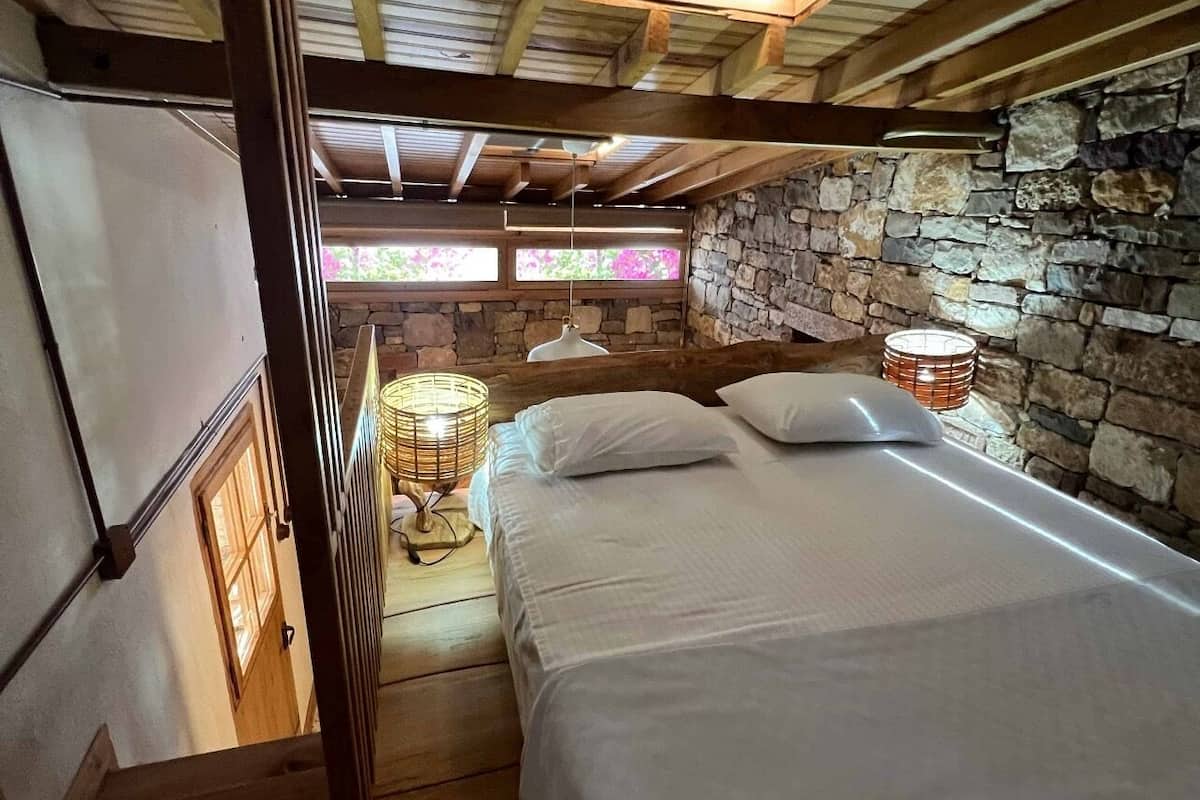 A bedroom with a stone wall, a bed, a hanging lamp, and a window with a view of greenery.
