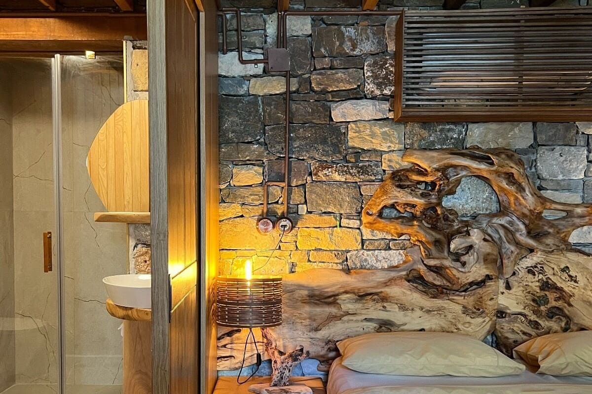 A bedroom with a stone wall, wooden beams, a bed, a nightstand, a lamp, and a glass shower enclosure.