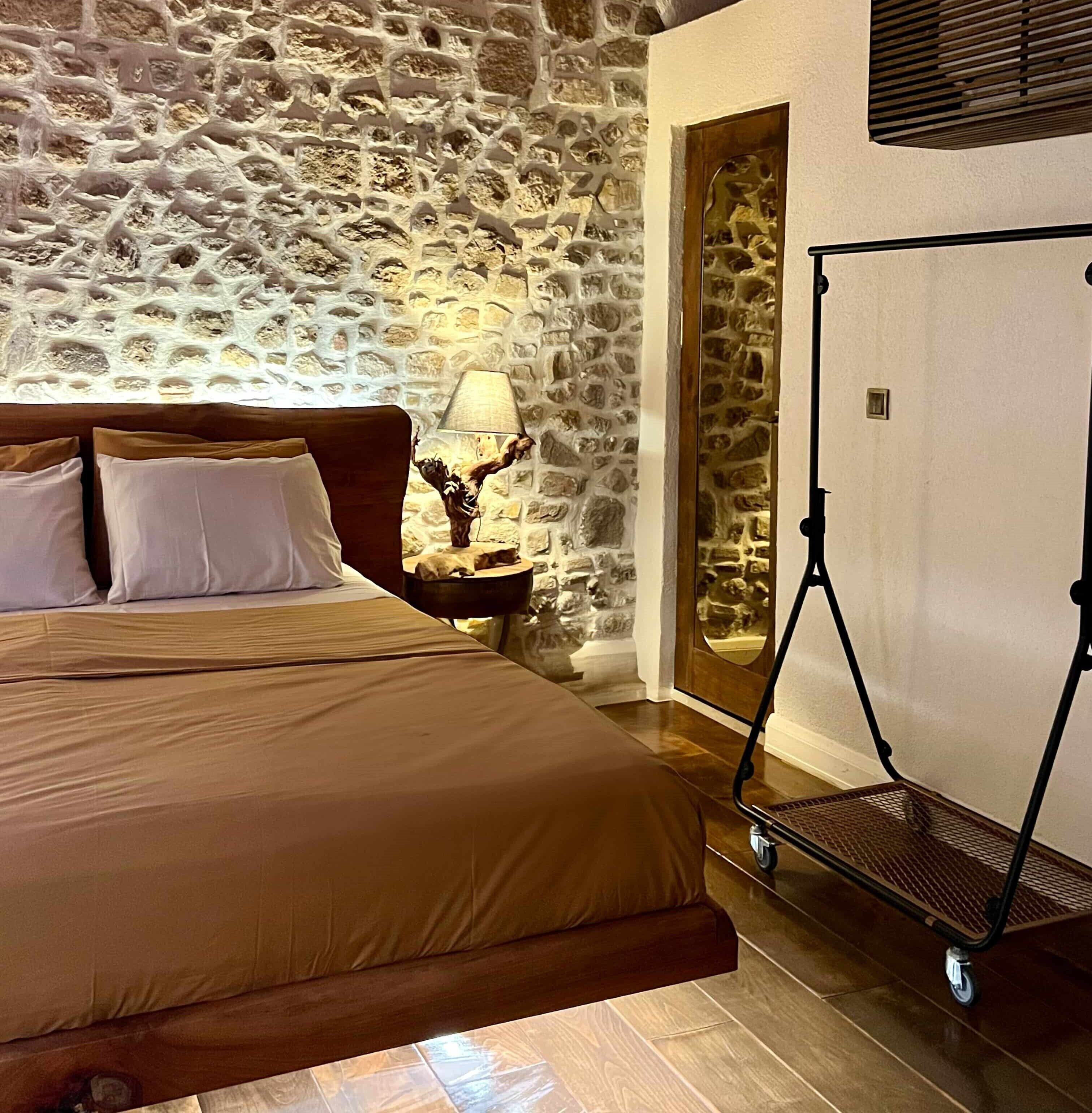 Un dormitorio con pared de piedra, una cama con cabecera, una mesita de noche con lámpara y un carrito con una pantalla.