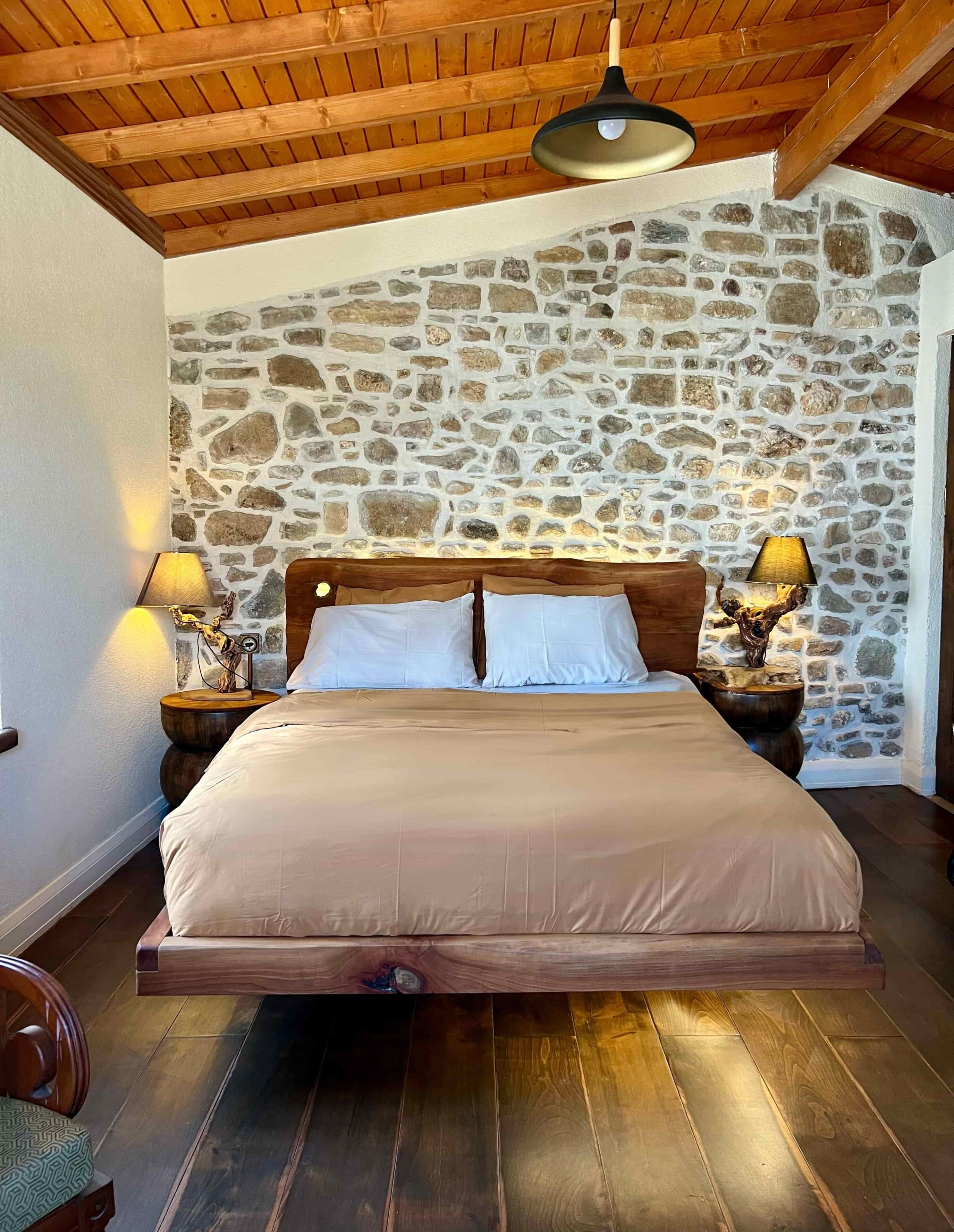 Un dormitorio con pared de piedra, vigas de madera en el techo, una cama grande, dos lámparas de noche y suelo de madera.