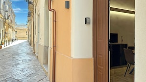 Uma rua estreita com entrada de prédio, uma porta e um pequeno espaço ao ar livre com mesas.