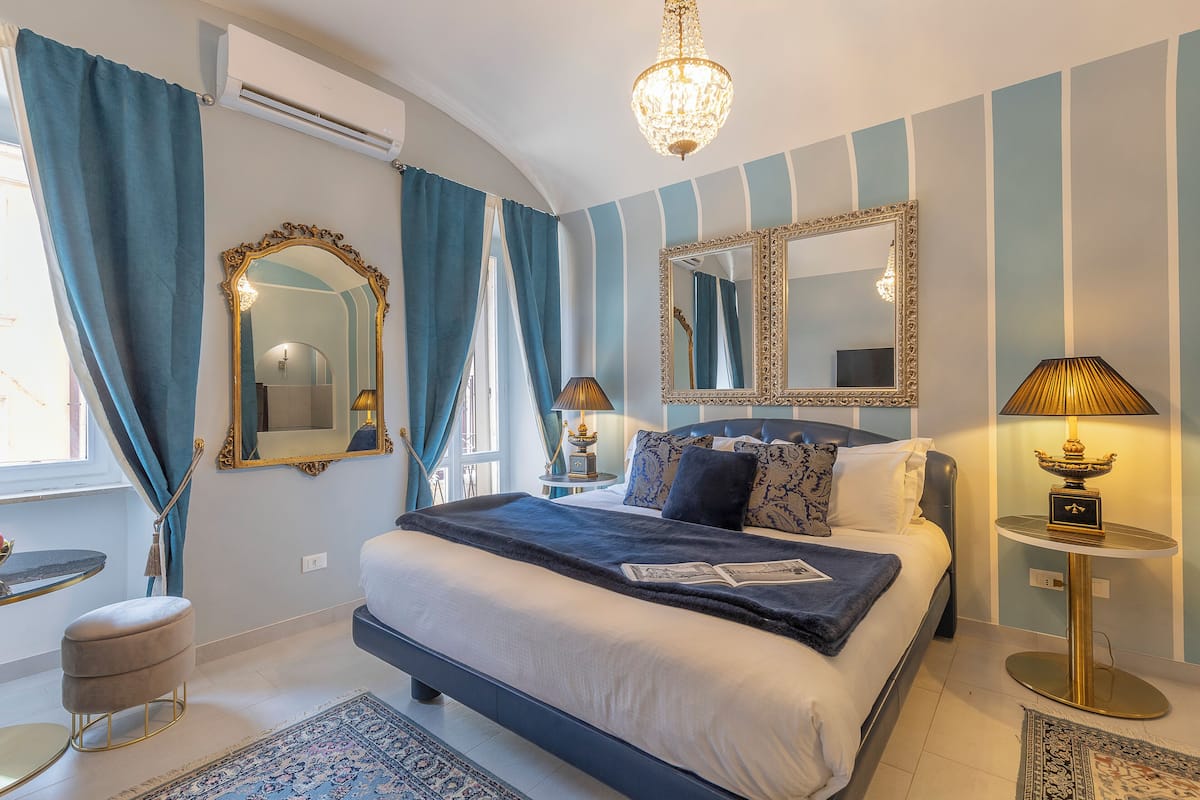 A bedroom with a large bed, a chandelier, a mirror, and a bedside table with a lamp.