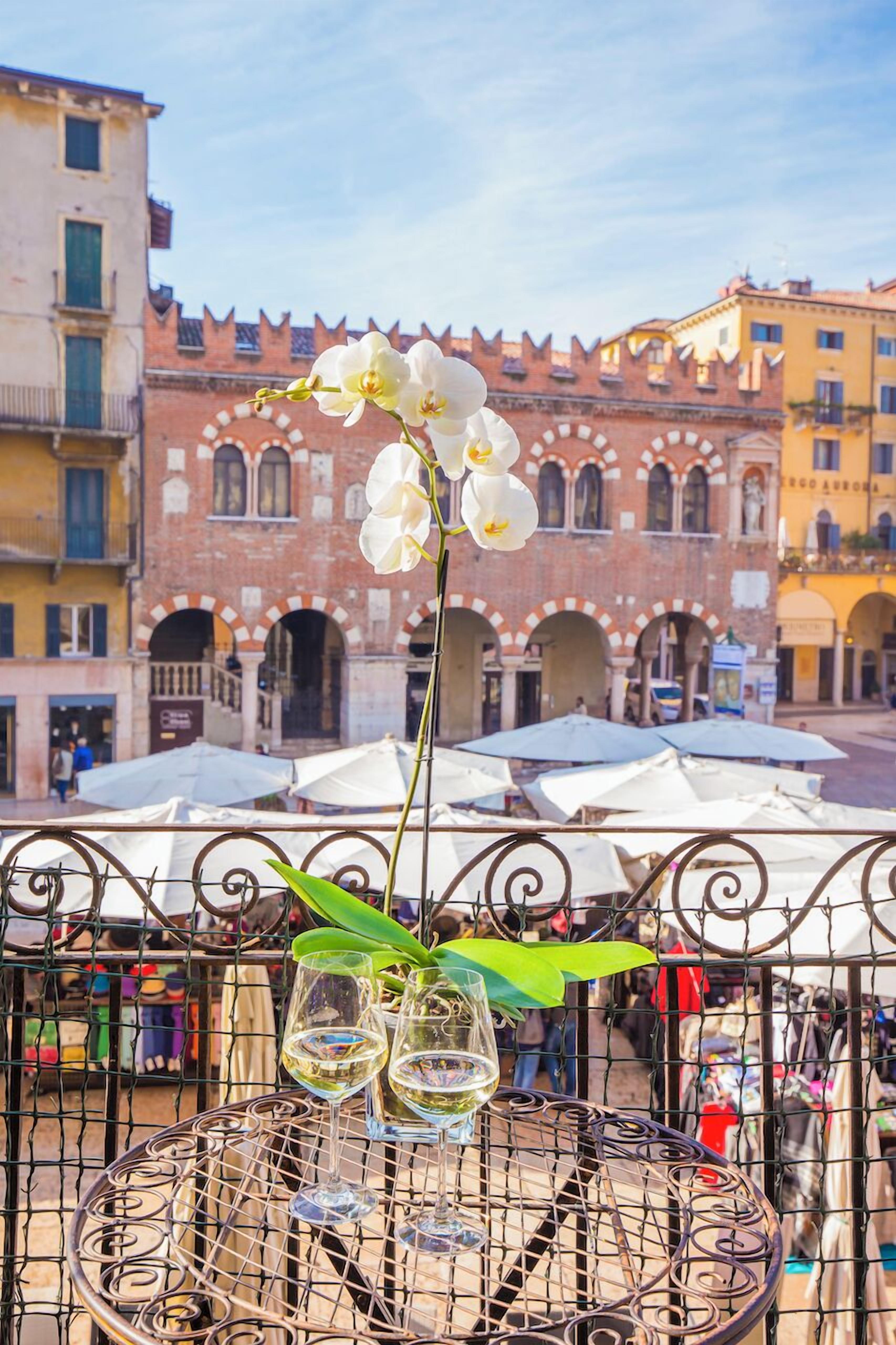 Une table avec deux verres à vin et un vase de fleurs blanches, le tout placé devant un bâtiment historique doté d’arcades et sous un ciel dégagé.