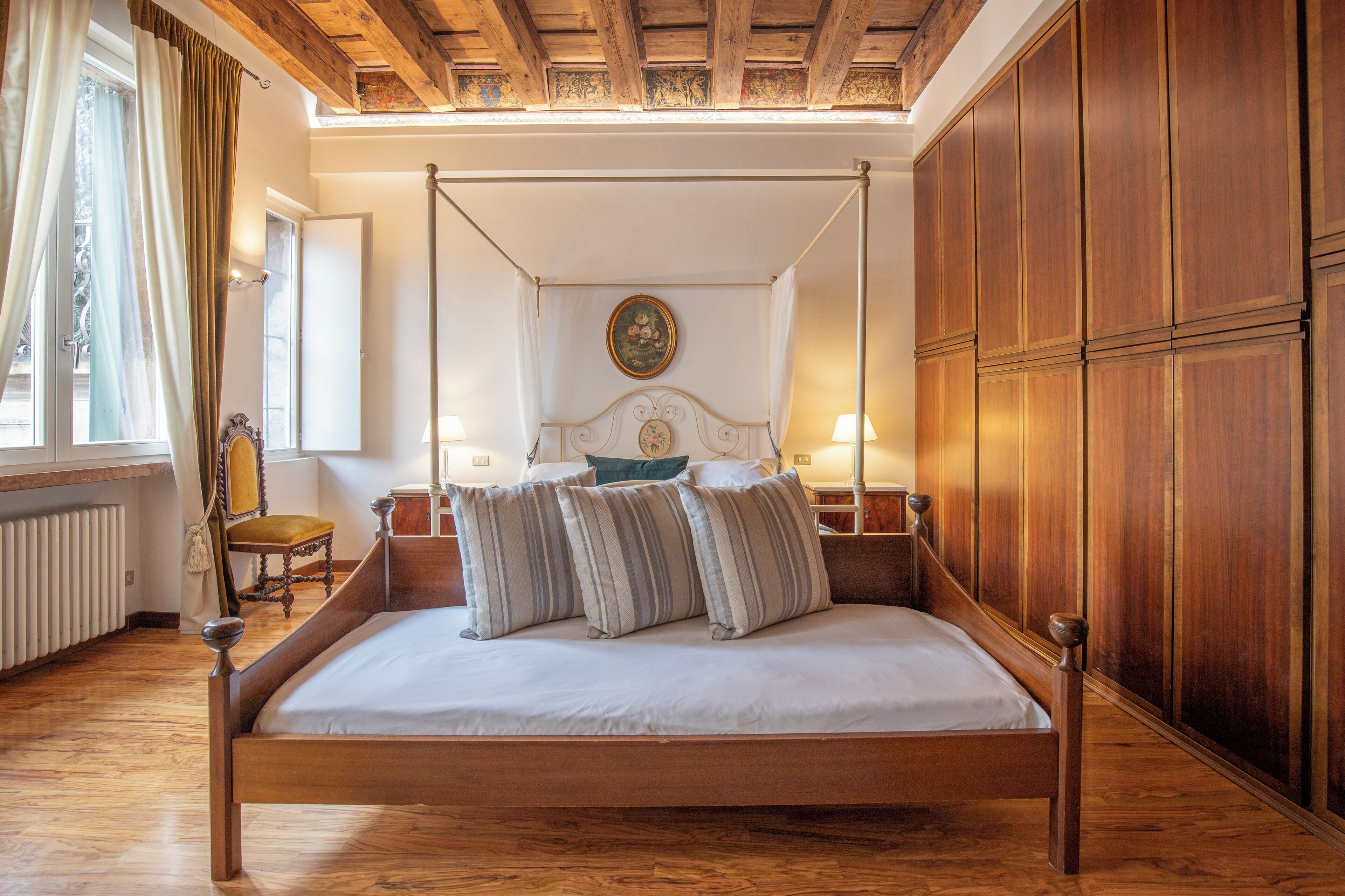 Un lit à baldaquin avec une structure en bois et une literie blanche, une armoire en bois sur la droite, une petite table avec une chaise, et une fenêtre avec des rideaux sur la gauche.