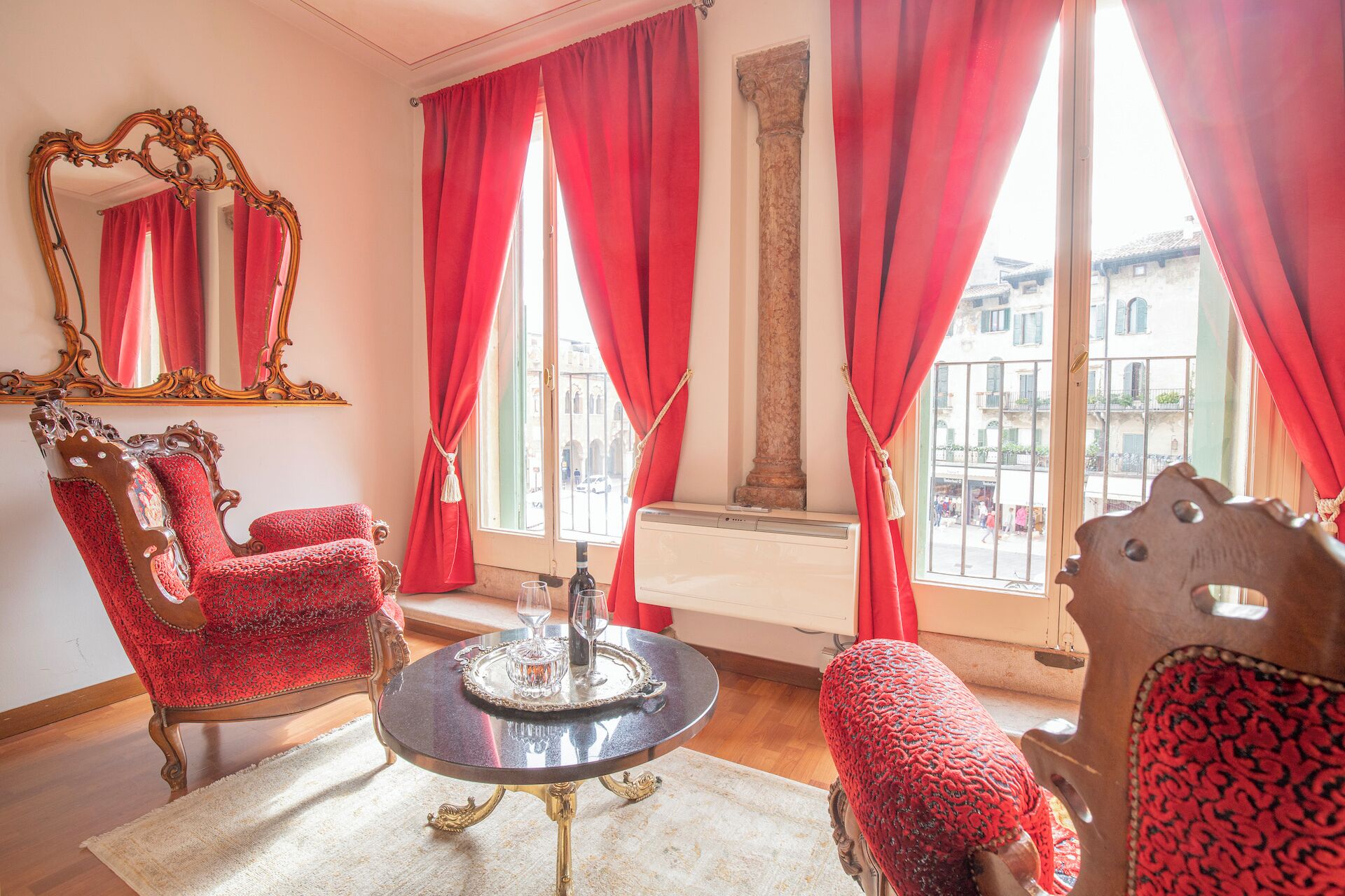 Una stanza con poltrone rosse decorate, un tavolo rotondo con vassoio, uno specchio grande e vista sugli edifici attraverso ampie finestre con tende rosse.