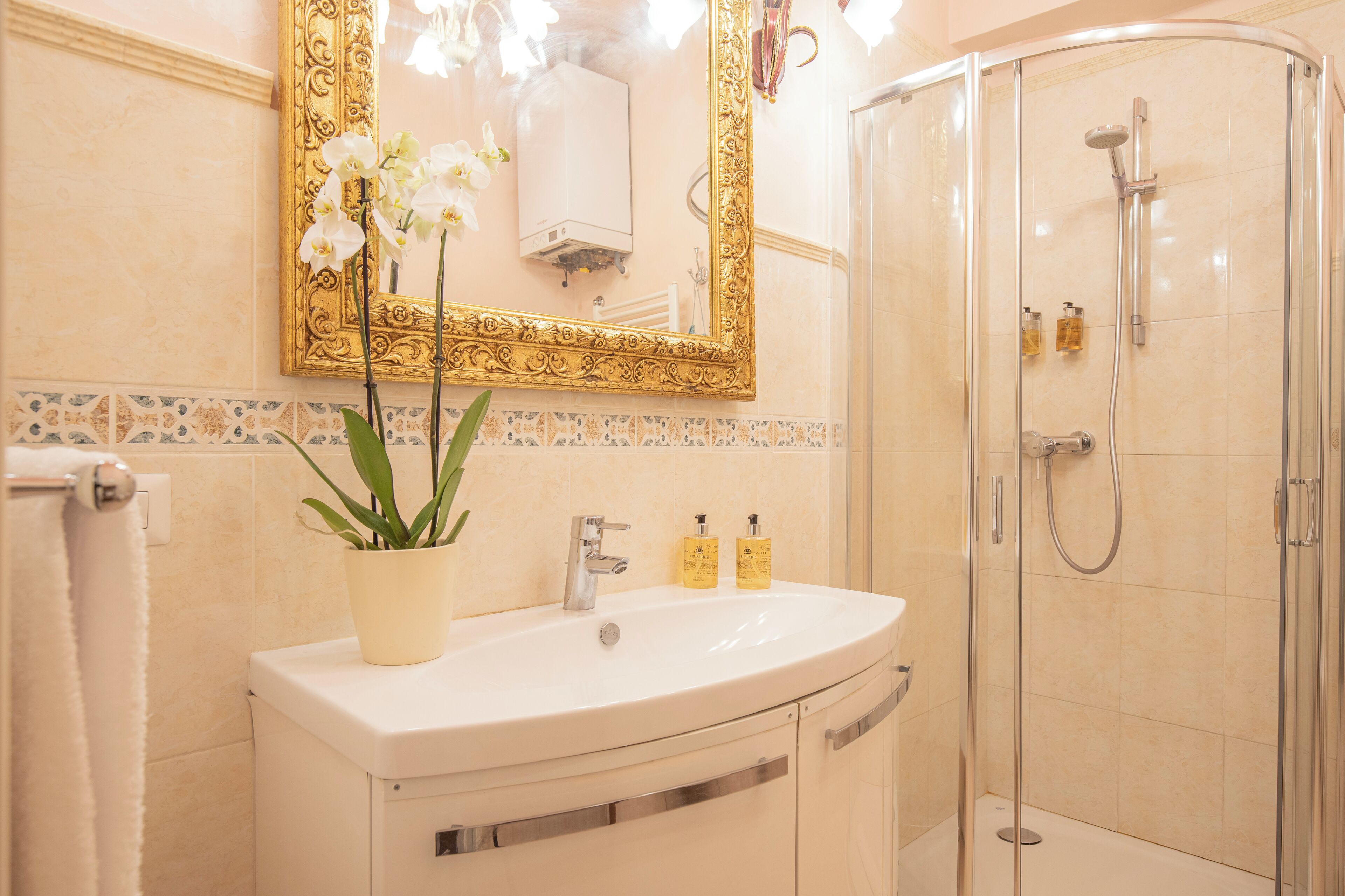 A bathroom with a white sink, a gold-framed mirror, a potted orchid, and a glass-enclosed shower.