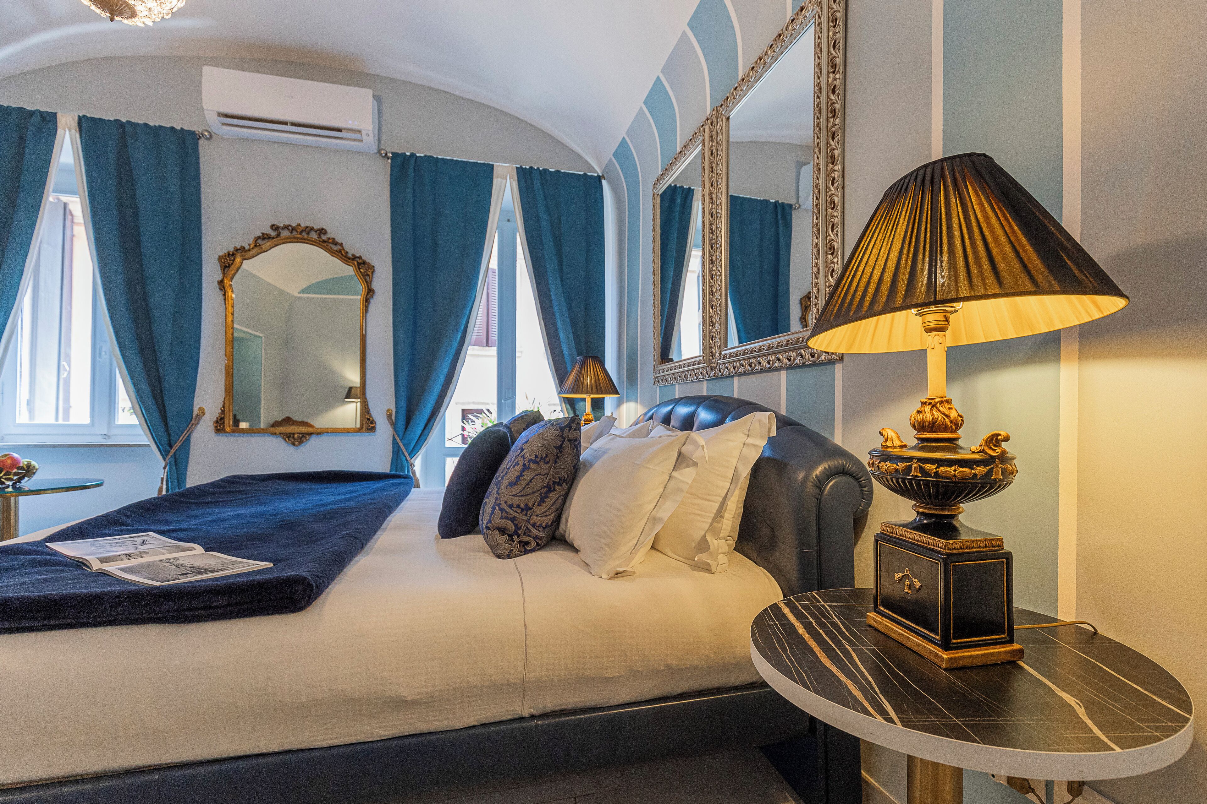 A hotel room with a bed, a round table with a lamp, a mirror, and blue curtains.