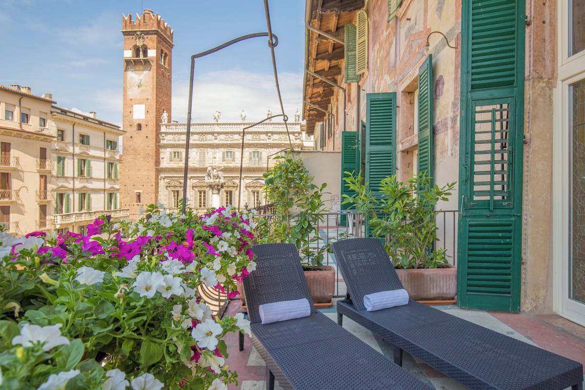 A balcony with lounge chairs, potted plants, and a view of a historic building with a tower.