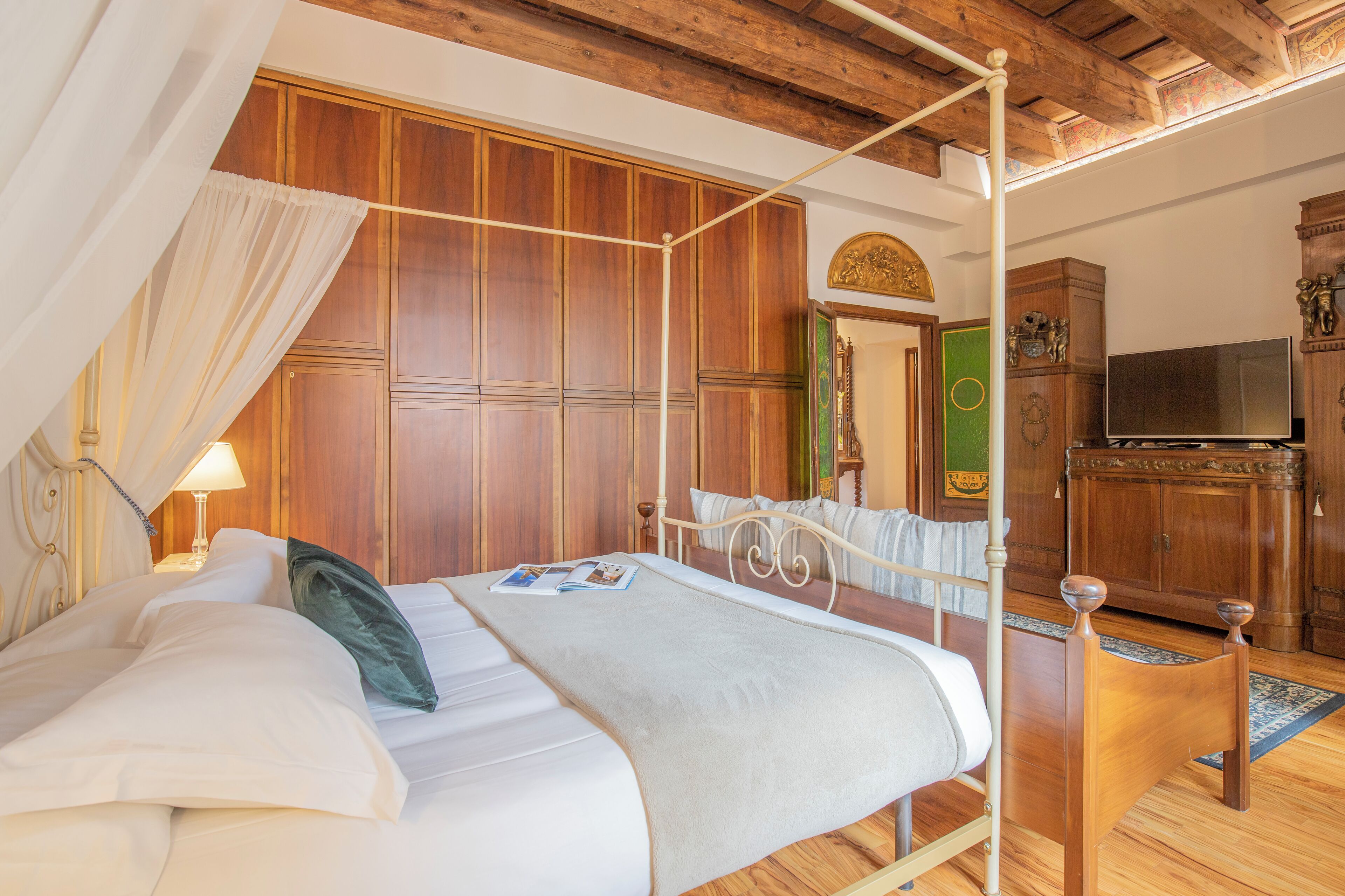 A four-poster bed with a canopy, wooden wardrobe, and a television in a room with wooden flooring and a green door.