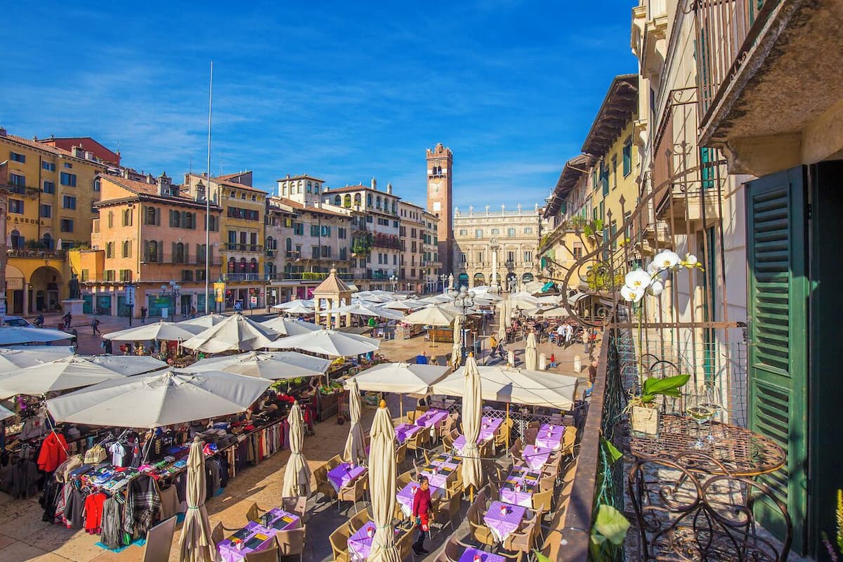 An outdoor market with stalls and tables under umbrellas, surrounded by historic buildings and a prominent tower.