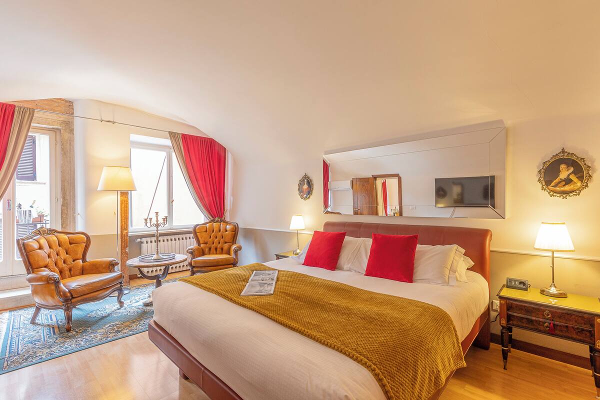 A hotel room with a large bed, two armchairs, a small table, a lamp, a clock, and a mirror.