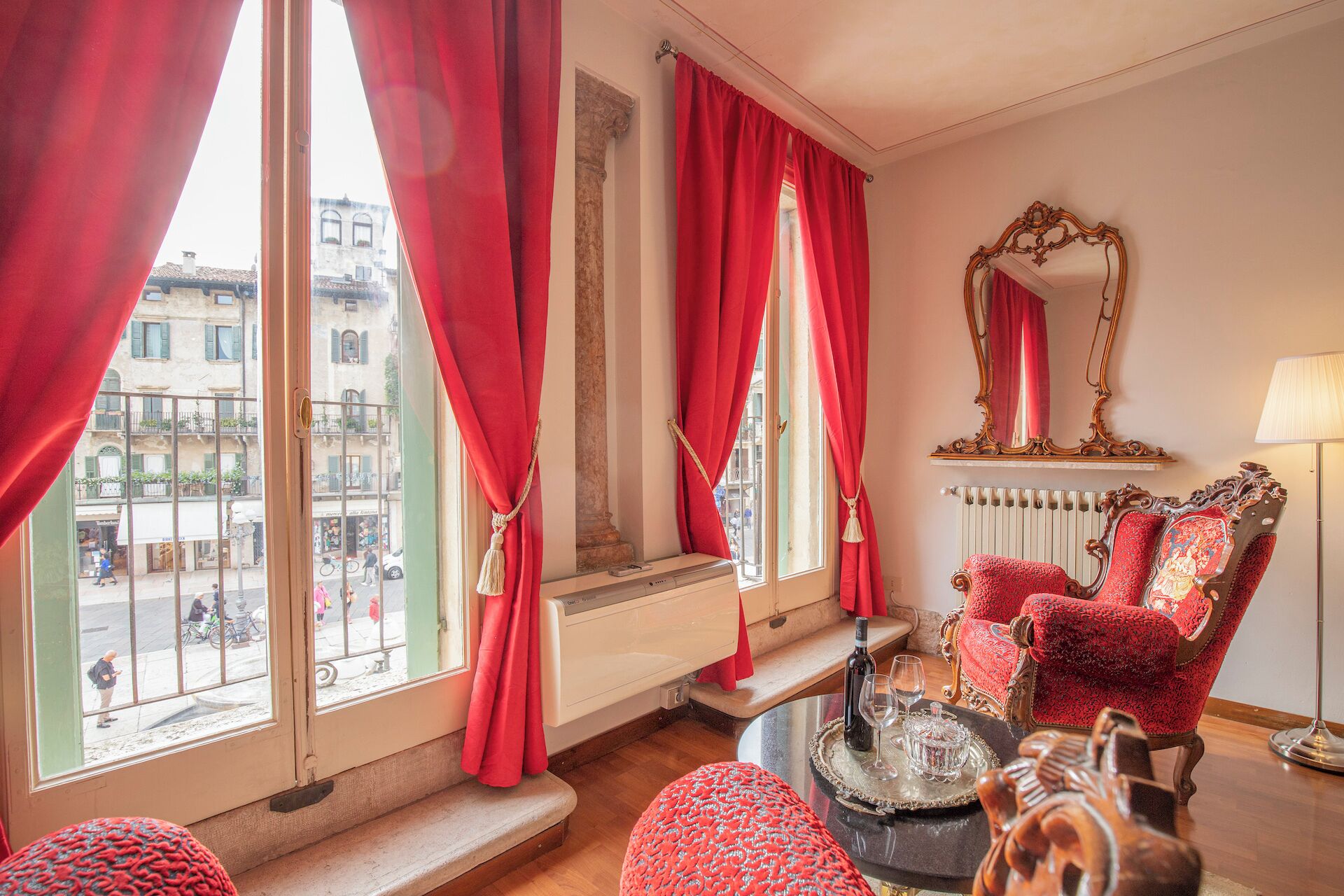 A room with large windows, red curtains, a classic chair, a table with a bottle and glasses, and a mirror on the wall.