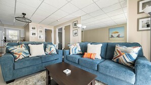 A living room with two blue sofas, a wooden coffee table, and a ceiling fan. - Oceanfront condos with 3 night minimum rentals and linens included! (Ocean City)