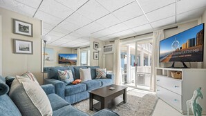 A modern living room with a blue sofa, a coffee table, and a television displaying a cityscape. - Oceanfront condos with 3 night minimum rentals and linens included! (Ocean City)
