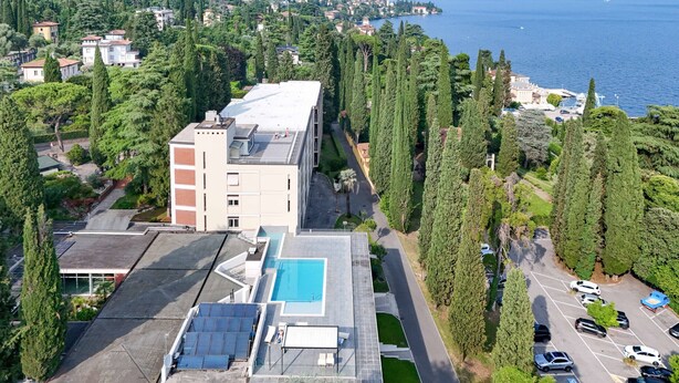 A modern building with a rooftop pool and solar panels, surrounded by tall trees and a parking lot. - MGH - Lake & Love Apartment (Gardone Riviera)