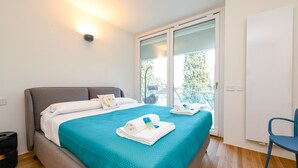 A modern hotel room with a large bed, a teal bedspread, and a view of greenery through a sliding glass door. - MGH - Lake & Love Apartment (Gardone Riviera)
