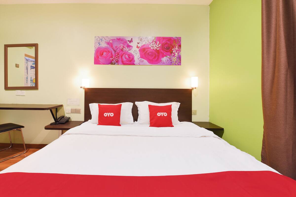 Een hotelkamer met een bed, twee rode kussens met 'OYO'-branding, een nachtkastje, een bureau met een stoel, en een schilderij van roze bloemen aan de muur.