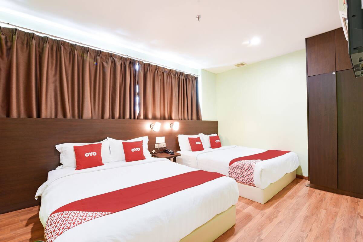 Een hotelkamer met twee bedden, elk voorzien van witte beddengoed en rode accenten, een houten hoofdbord, een bruin gordijn en een houten vloer.