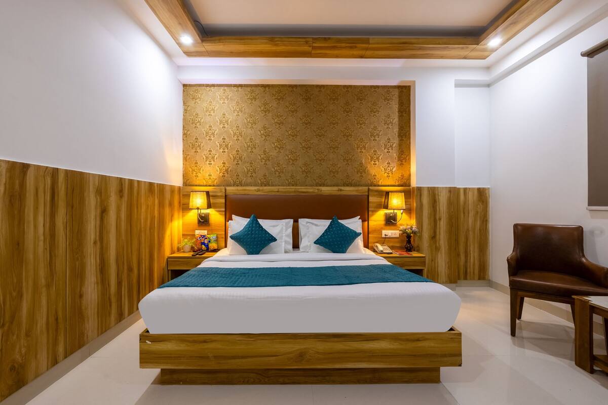 Ein Hotelzimmer mit einem Holzbettgestell, einem großen Bett mit weißer Bettwäsche und blauen Akzenten, zwei Nachttischen mit Lampen, einem Holzstuhl und einer Wand mit gemustertem Tapetenpapier.