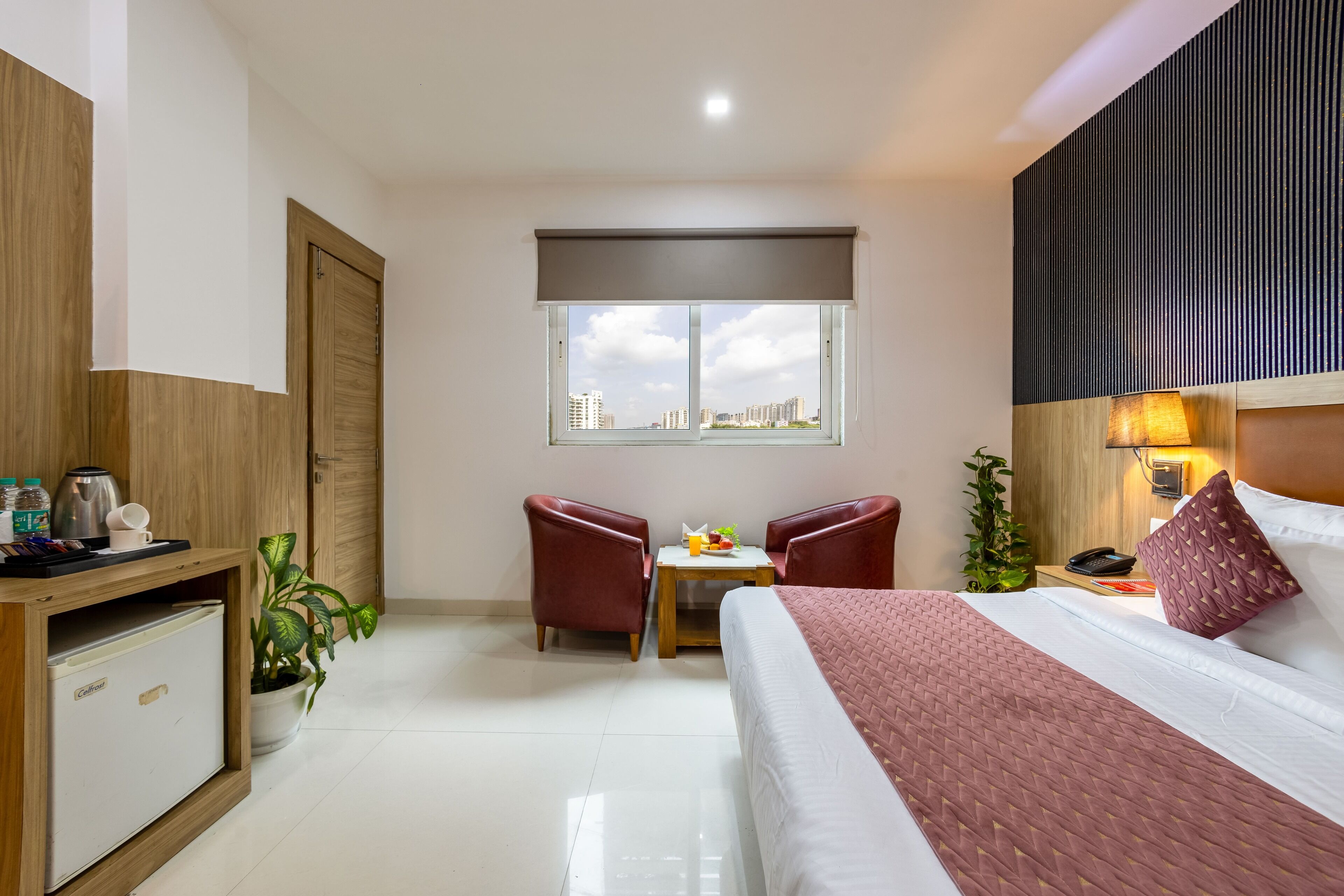 A hotel room with a bed, a desk with a computer, a chair, a small table with a plant, and a window with a city view.