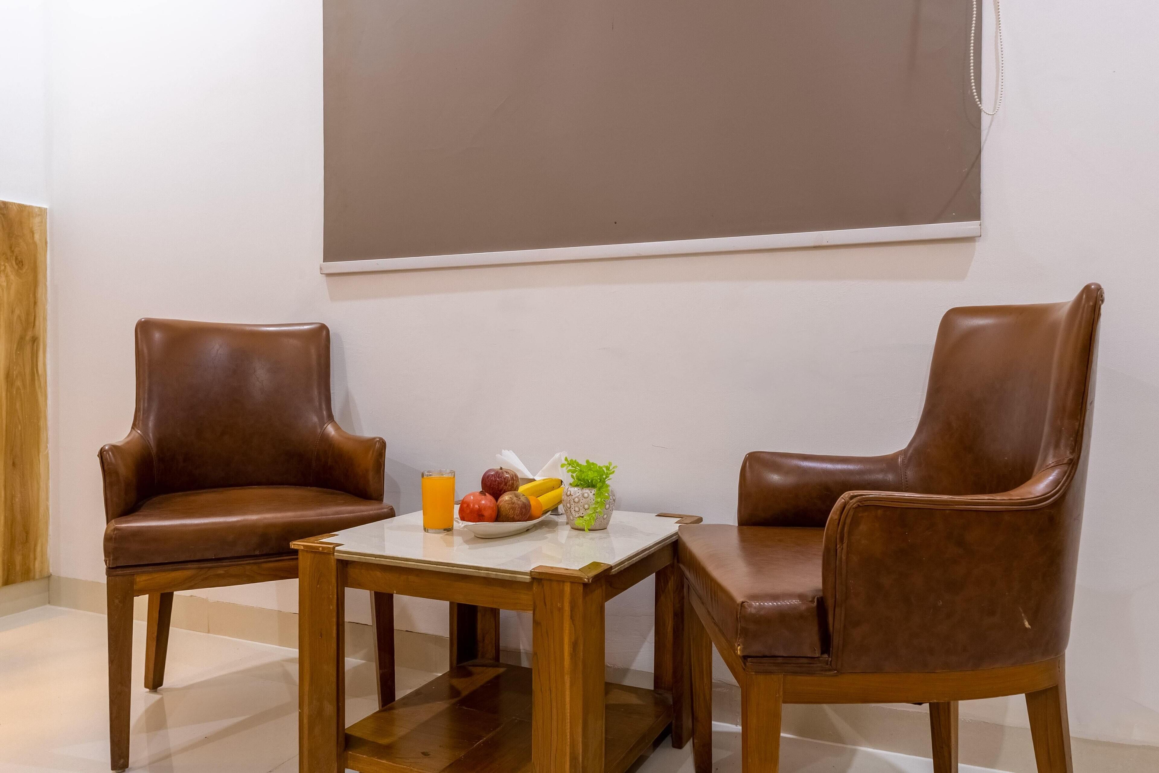 Two brown leather chairs, a wooden table, and a fruit bowl with a drink on a white table in a room with a large window and a plain wall.
