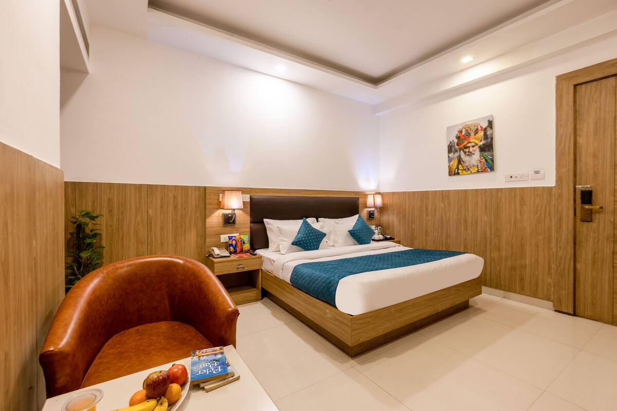 Ein Hotelzimmer mit Bettgestell aus Holz, einem Nachttisch mit Lampe, einem braunen Ledersessel und einem kleinen Tisch mit Früchten und einem Buch.