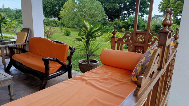 A balcony with a wooden bench and chairs, a potted plant, and a view of a lush garden. - Spacious 5-Bedroom Homestay Villa in Charming Malindi  WiFi Included (Malindi)