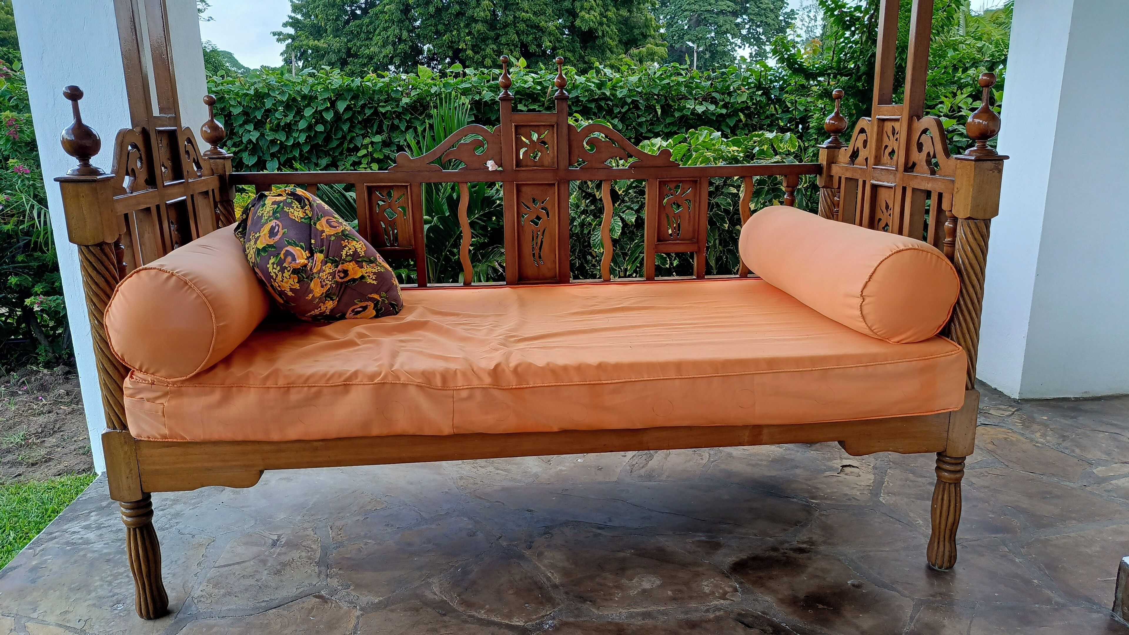 Un banc en bois agrémenté de coussins orange et d’un coussin à motifs floraux, placé sur une terrasse en pierre avec un mur blanc et de la verdure en arrière-plan.