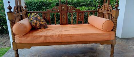 Un banc en bois agrémenté de coussins orange et d’un coussin à motifs floraux, placé sur une terrasse en pierre avec un mur blanc et de la verdure en arrière-plan.
