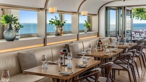 Un restaurante con vista al océano, que cuenta con una barra larga, asientos acolchados y plantas en macetas.