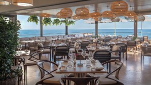 Un restaurante con mesas y sillas dispuestas para comer, amplias ventanas con vistas al mar y lámparas colgantes de mimbre.