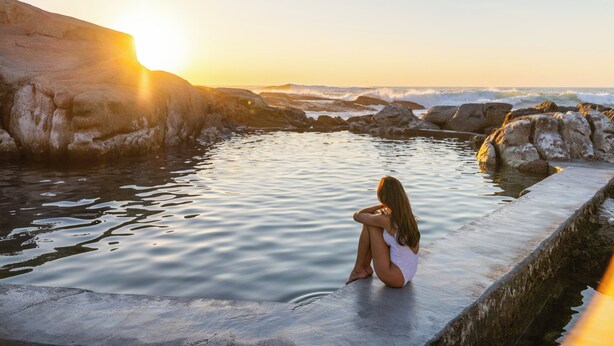 Una mujer sentada junto a un cuerpo de agua tranquilo, con una costa rocosa y un atardecer de fondo.