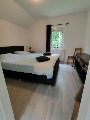 A bedroom with a bed, a window with curtains, a radiator, a chair, and a small table with a lamp. - Vacation home Knabbel & Babbel (Lochem)
