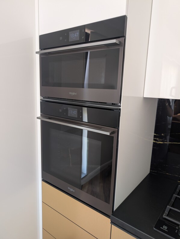 A stainless steel Whirlpool oven with a digital clock displaying 13:47, integrated into a modern kitchen with white cabinetry and a black countertop.