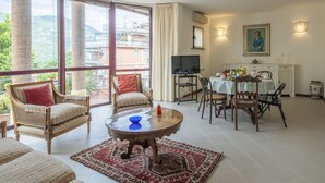 Un salon spacieux avec une grande fenêtre, un coin repas avec une table dressée pour deux personnes et un téléviseur posé sur un meuble.