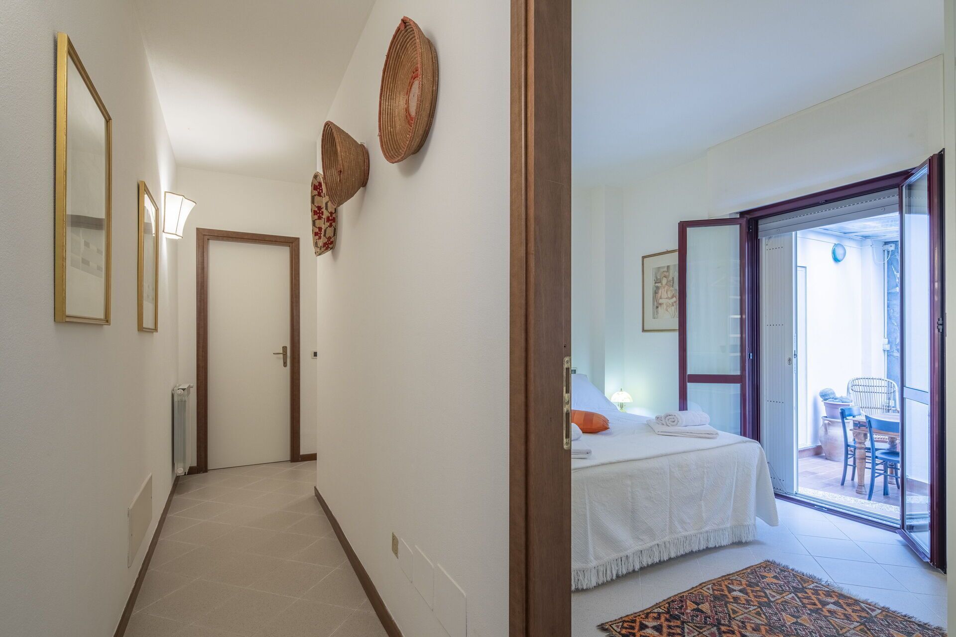Corredor que dá para um quarto com cama, uma mesa pequena e uma cadeira. Há duas portas, uma para o quarto e outra para uma varanda.