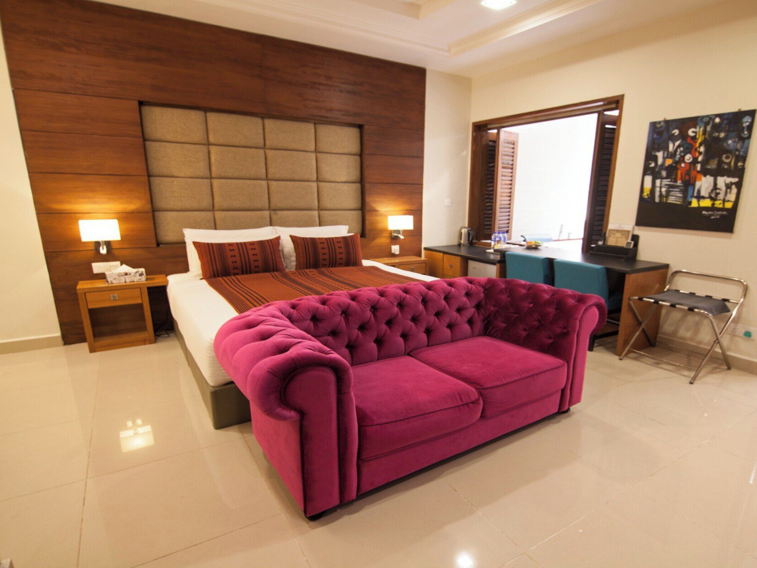 A modern hotel room with a large bed, a tufted pink sofa, a wooden headboard, a desk, and a large window.