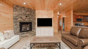 Un salón acogedor con chimenea de piedra, televisor de pantalla plana, mueble blanco para equipo multimedia, sofá de cuero marrón y una mesa de centro de madera.