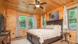Un dormitorio de cabaña de madera con cama, ventilador de techo y ventanas amplias.