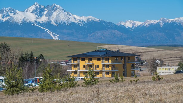 Ein gelbes Gebäude mit mehreren Balkonen, umgeben von einer Wiese und Bergen im Hintergrund.