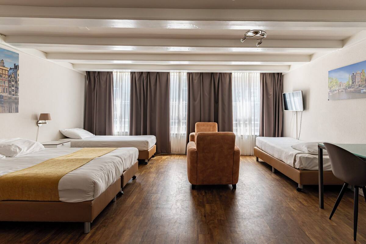 Habitación de hotel con dos camas, un escritorio, una silla, un televisor y ventanas grandes con cortinas.